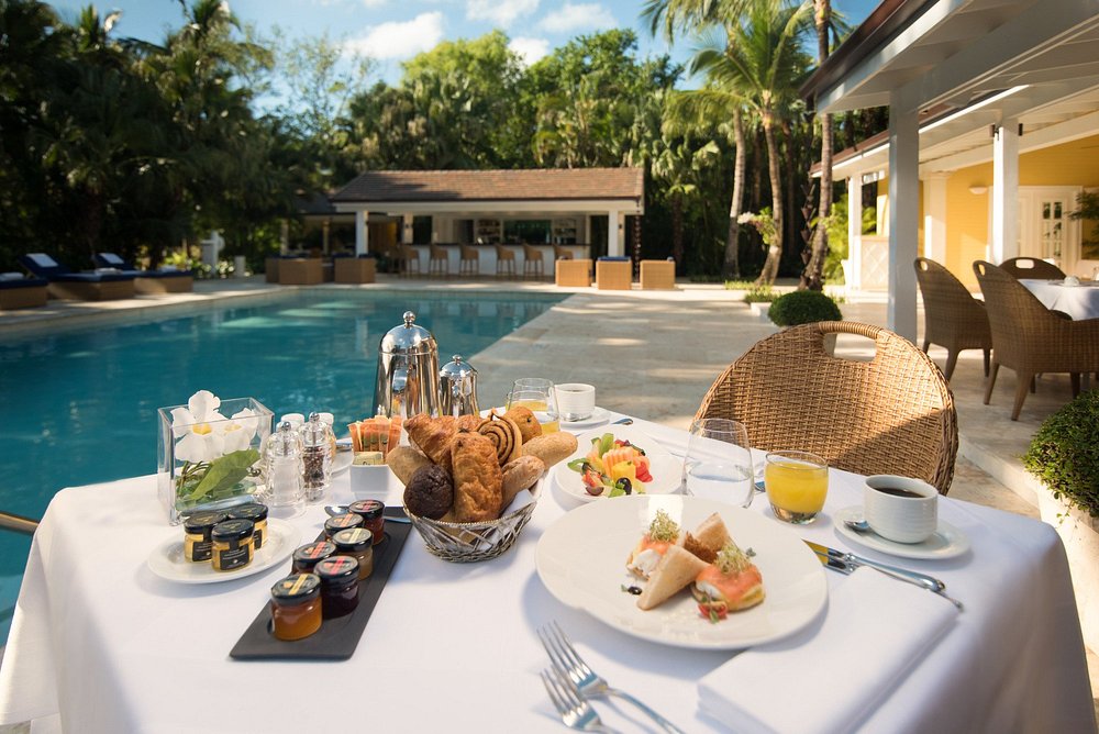 A luxurious breakfast spread on a table beside a swimming pool, featuring a variety of pastries, fruits, jams, and beverages with a tropical garden backdrop.