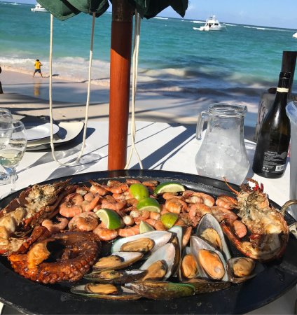 A seafood platter featuring shrimp, mussels, and octopus, served on a table with a beach view, including boats in the background and a person walking on the shore.