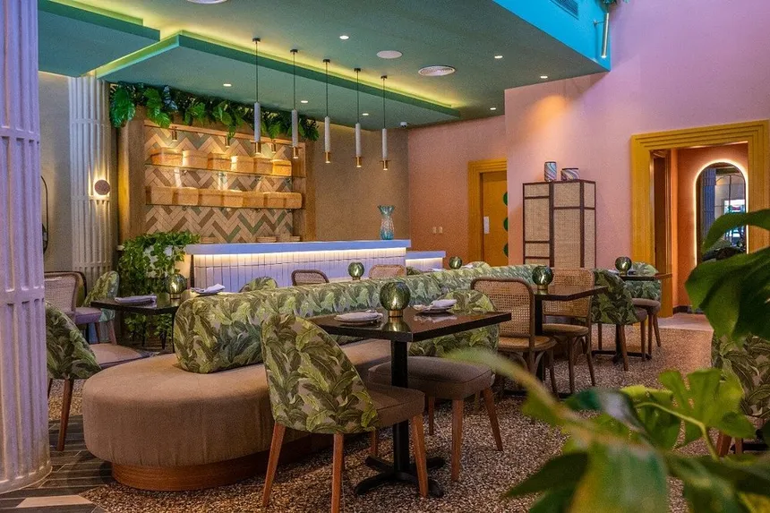 Interior view of a modern restaurant featuring lush greenery, stylish seating, and a cozy ambiance. The design includes nuanced lighting, tropical-patterned upholstery, and decorative elements on the walls.