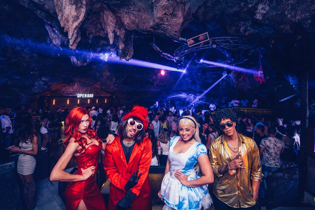 imagine-cave-nightclub Four people in colorful costumes posing at a lively nightclub with an open bar sign and blue stage lights in a cave-like setting