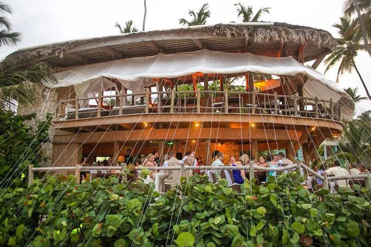 A beachfront restaurant with a thatched roof and wooden structure, surrounded by palm trees and lush greenery. The outdoor dining area is bustling with people enjoying their meals.