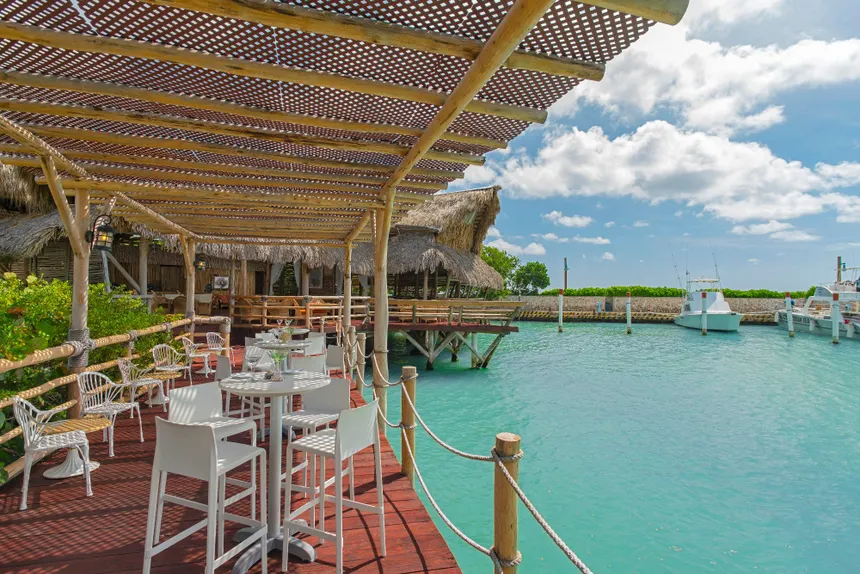 A waterfront dining area with white tables and chairs under a bamboo canopy, overlooking turquoise water and a boat dock.