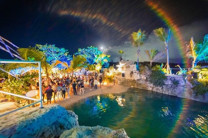 maroca-puntacana Nighttime beach scene with crowd, illuminated palm trees, rocky pool, and a rainbow halo in the sky