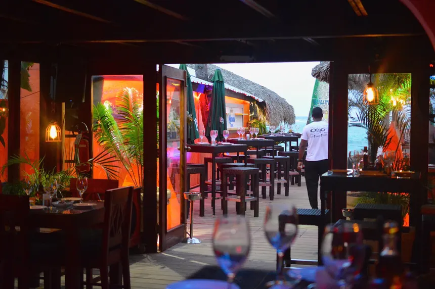 Interior view of a tropical restaurant with wooden tables, glassware set for dining, and a server walking towards the outdoor area, featuring decorated lighting and palm plants.
